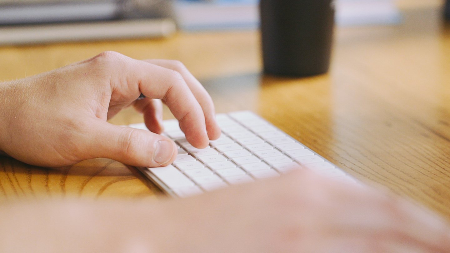 Mani che digitano su una tastiera bianca su una scrivania di legno.